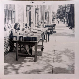 Volunteers and staff sit at tables lined with books at a book sale in August 1965.